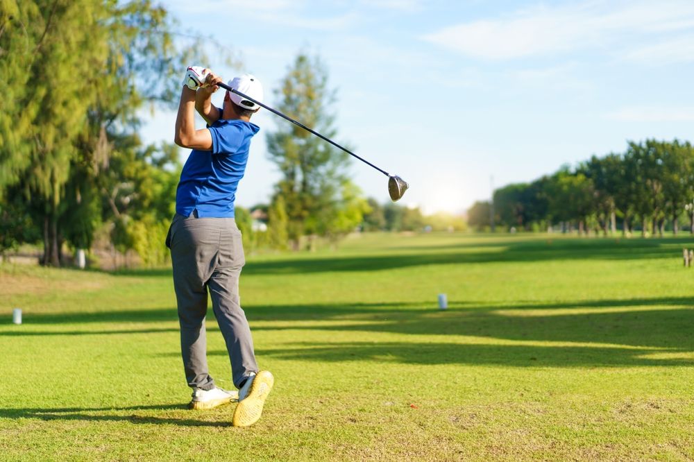 Golfer swings club on green course; blue shirt, gray pants, sunny day.
