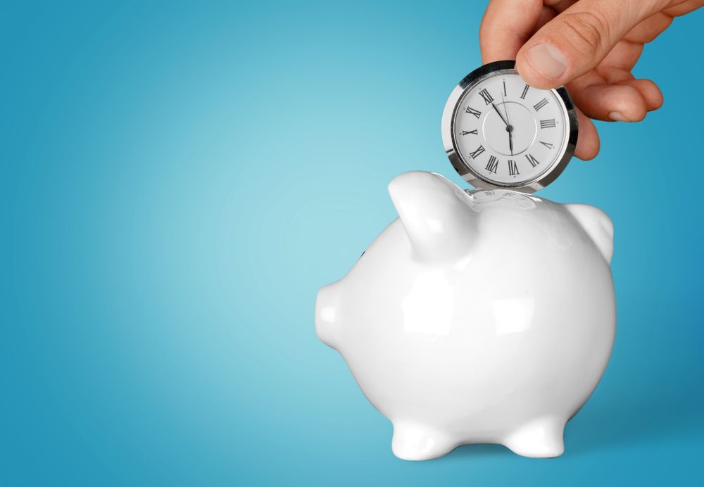 Hand placing a clock into a piggy bank, implying saving time or time as a valuable resource.