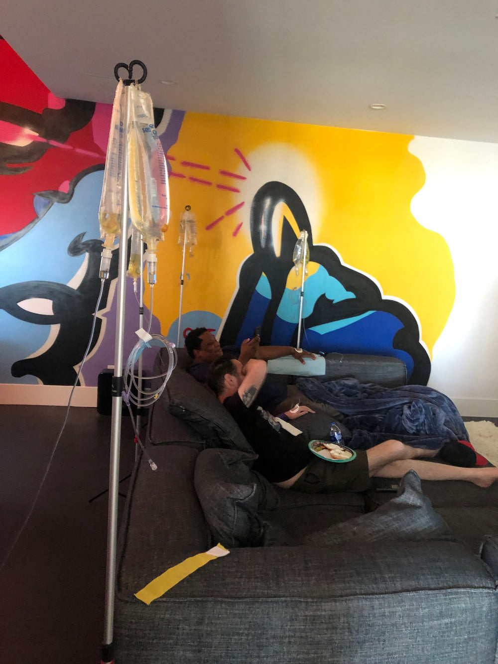 Person on couch receiving IV fluids, colorful mural backdrop.