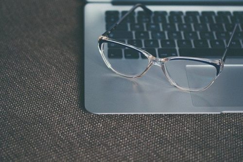 Clear-framed glasses rest on a laptop keyboard. The laptop is open on a textured, gray surface.