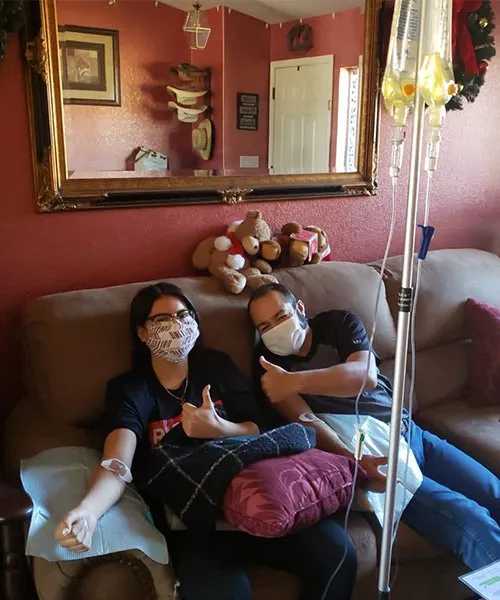 Two people giving thumbs up while receiving IV drips on a sofa. Both wear masks, setting is a home.