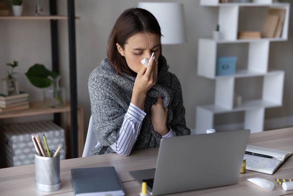 Woman with a cold sneezes into a tissue while working on a laptop at a desk.