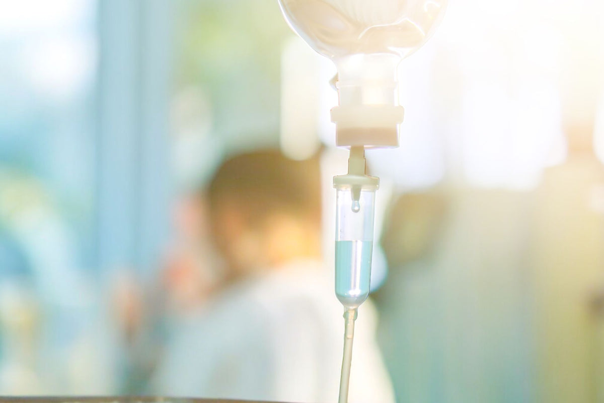 IV drip bag dispensing fluids, blurred background of a person in a medical setting.