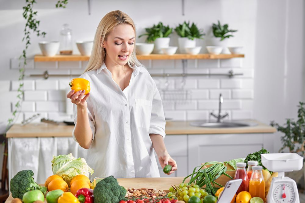Blonde woman in kitchen holds yellow pepper, surrounded by produce. Counter, shelves, sink in background.