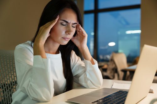 Woman with a headache, rubbing her temples, sitting at a desk with a laptop, in an office at night.