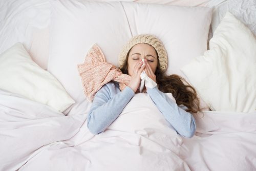 Woman in bed blowing nose, wearing hat and scarf; appears ill.