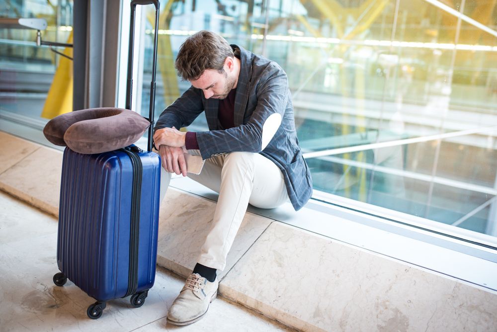 Man resting by a window at an airport, head down, suitcase and neck pillow nearby.