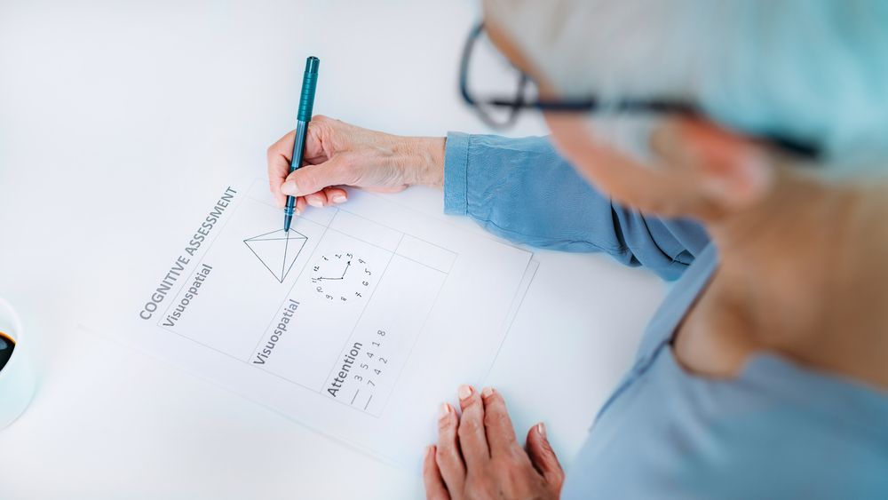 Person writing diagrams on white paper with a pen, wearing glasses and a blue top.