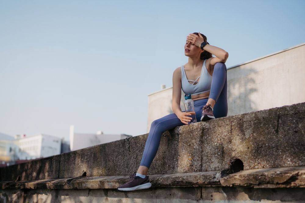 Woman in workout clothes sitting on a concrete ledge, resting hand on forehead, looking upward.