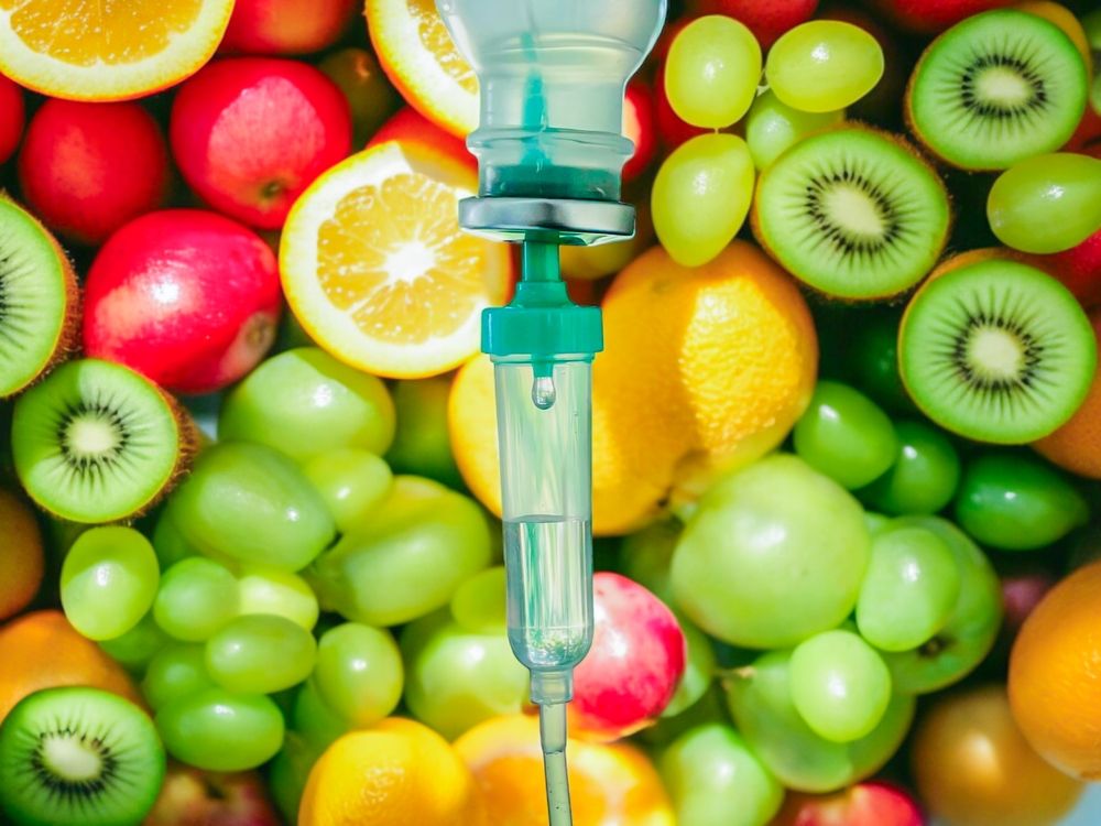 IV drip with green tubing against a background of fresh fruit including oranges, kiwi, and grapes.
