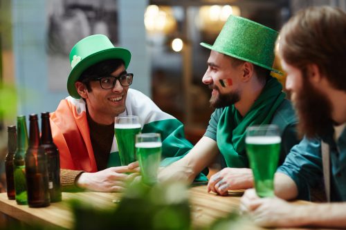 Three people in a bar celebrating St. Patrick's Day with green beer and hats; one is wrapped in an Irish flag.