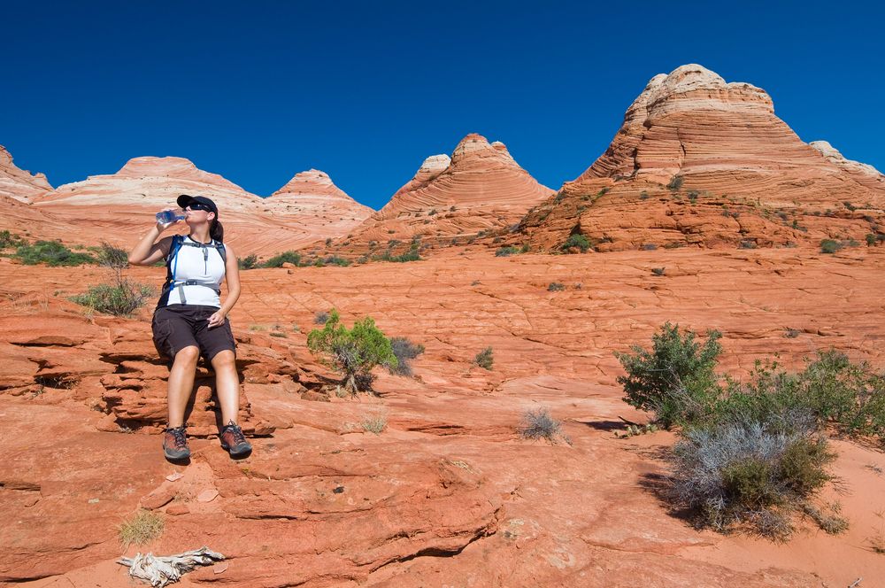 Woman hiking in a desert, drinking water, red rock formations, blue sky.