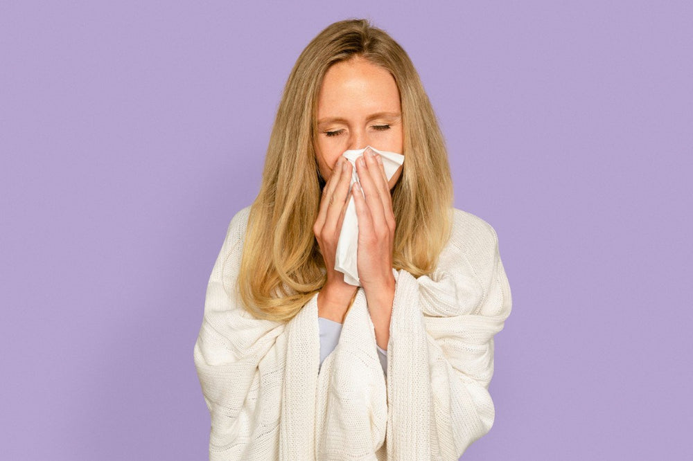 Woman wrapped in blanket blowing nose with tissue against a purple background.