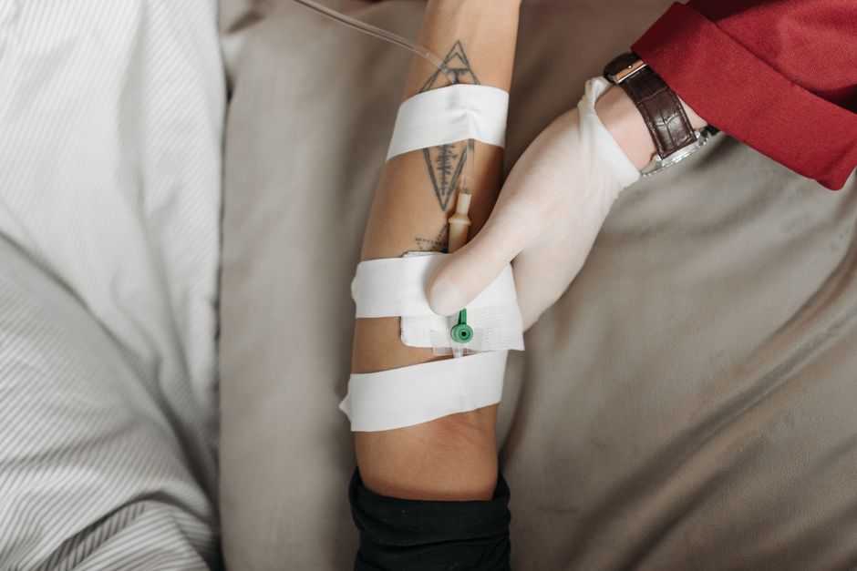 Arm with IV line secured by white tape, being tended to by gloved hand wearing watch.