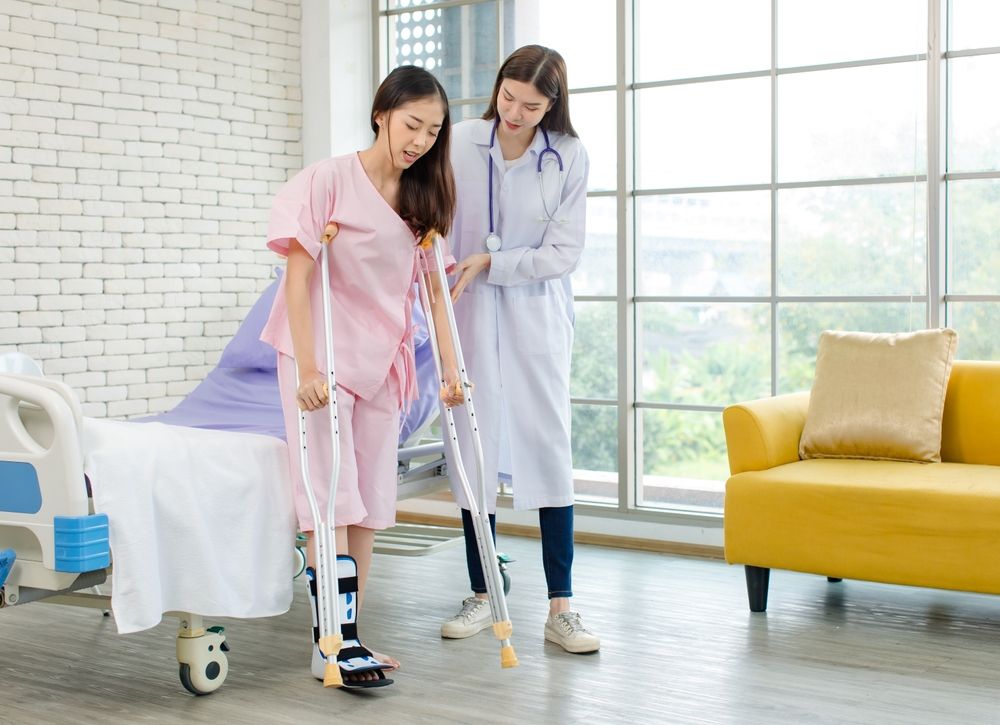 Woman with a leg brace using crutches, assisted by a doctor in a hospital room.