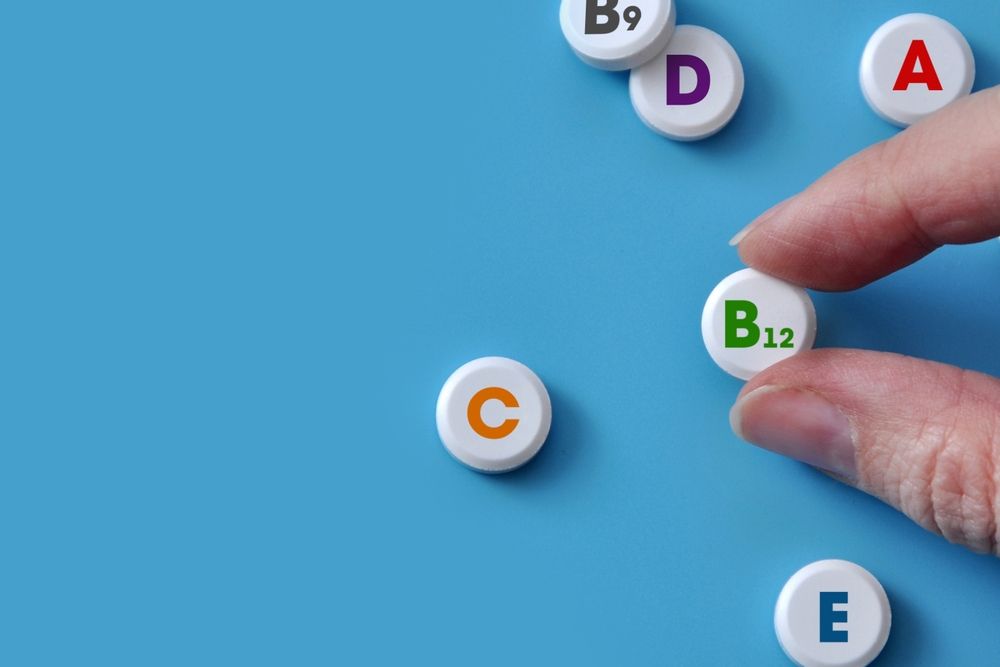 Hand picking a B12 vitamin from a scattered collection of vitamins labeled A, B9, C, D, and E on a blue surface.
