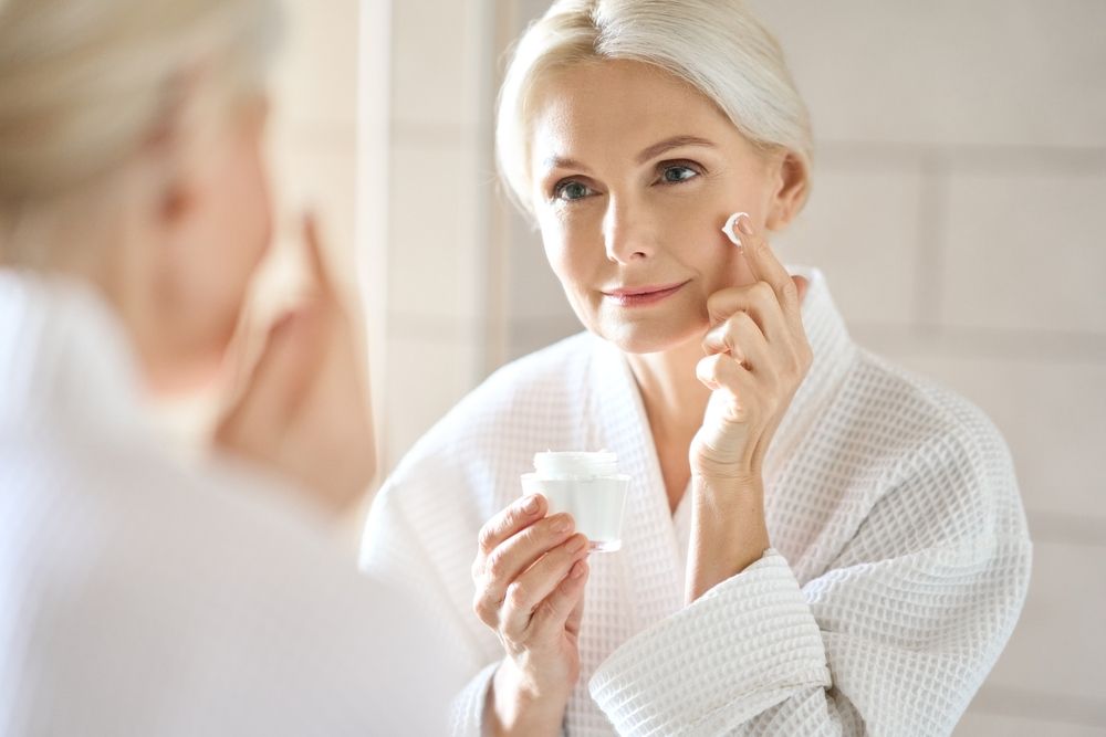Woman in a white robe applying cream to her face in front of a mirror.