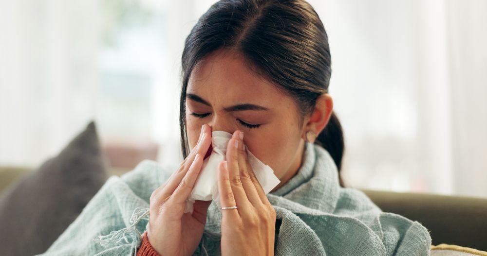 Woman blowing her nose with a tissue, wrapped in a blanket on a sofa.