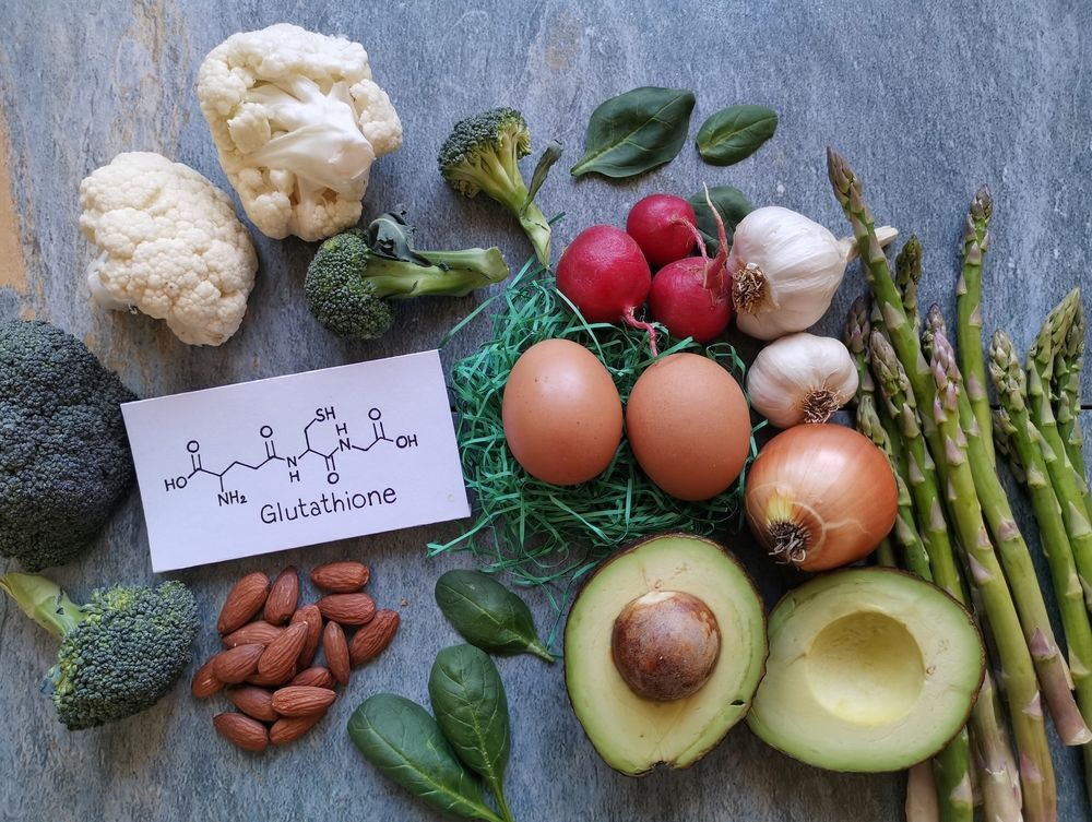 Vegetables and eggs arranged on a surface: broccoli, cauliflower, radishes, garlic, onion, asparagus, avocado, almonds, and eggs.