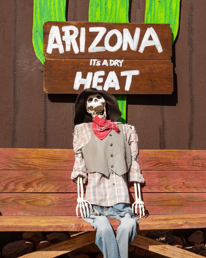 Skeleton dressed as a cowboy sitting on a bench beneath a sign that says