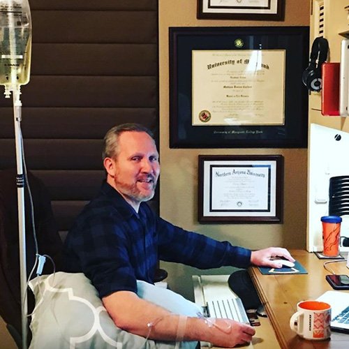 Man receiving IV fluids at desk, typing on keyboard. Wall with diplomas and headphones.