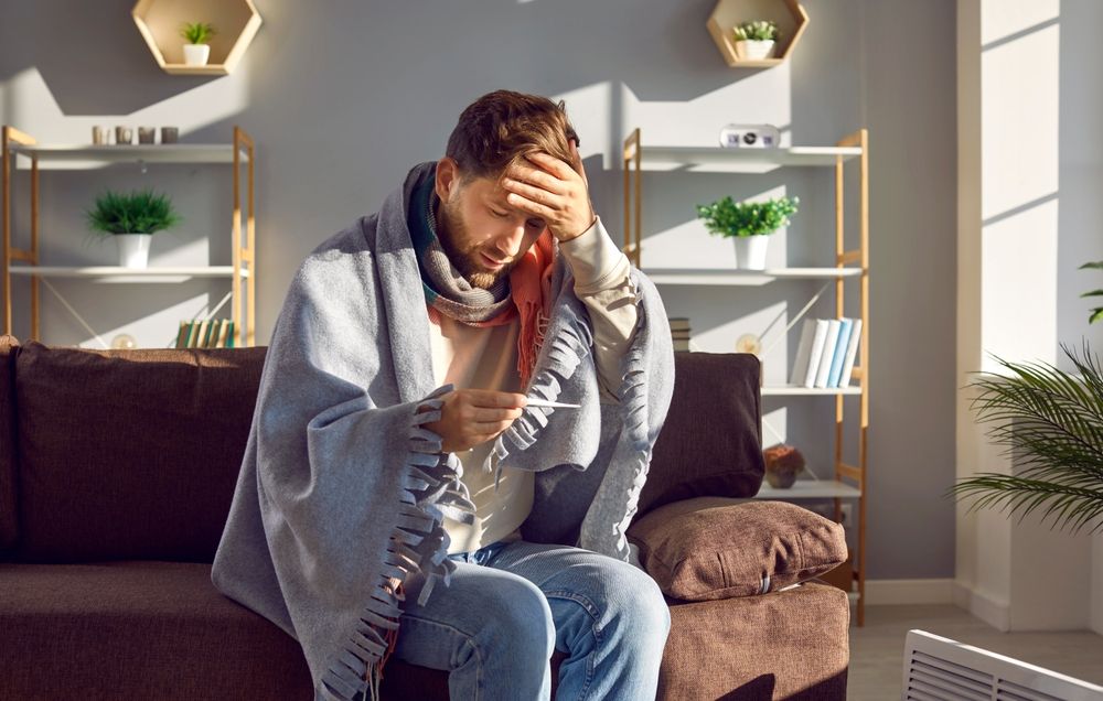 Man wrapped in blanket, checking a thermometer on a couch. Living room setting.