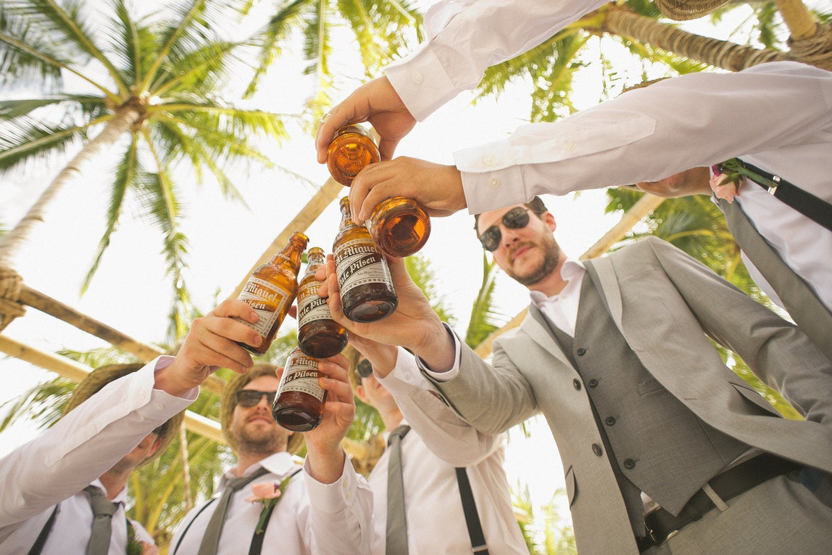 Men in suits toasting with beer bottles under palm trees.