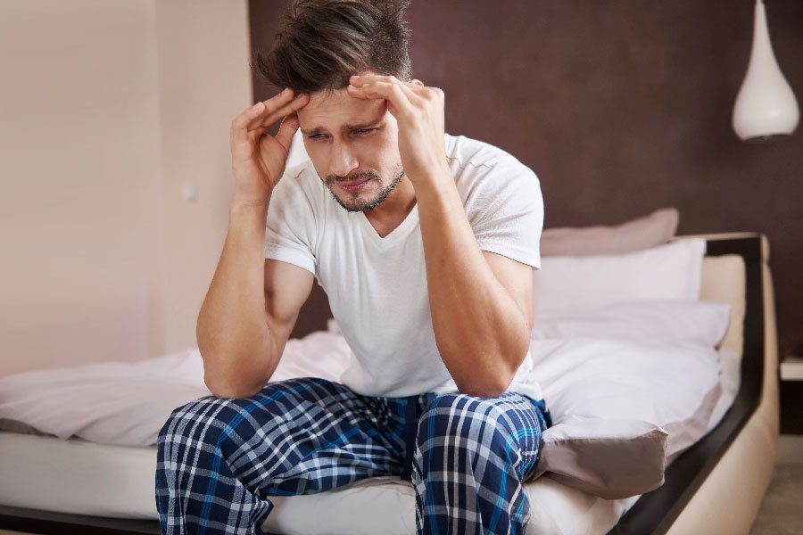 Man sitting on bed with hands on head, looking distressed.