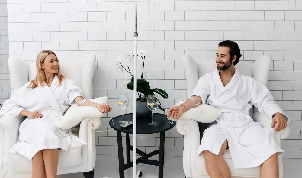 Two people in white robes receive IV infusions in a spa-like setting.