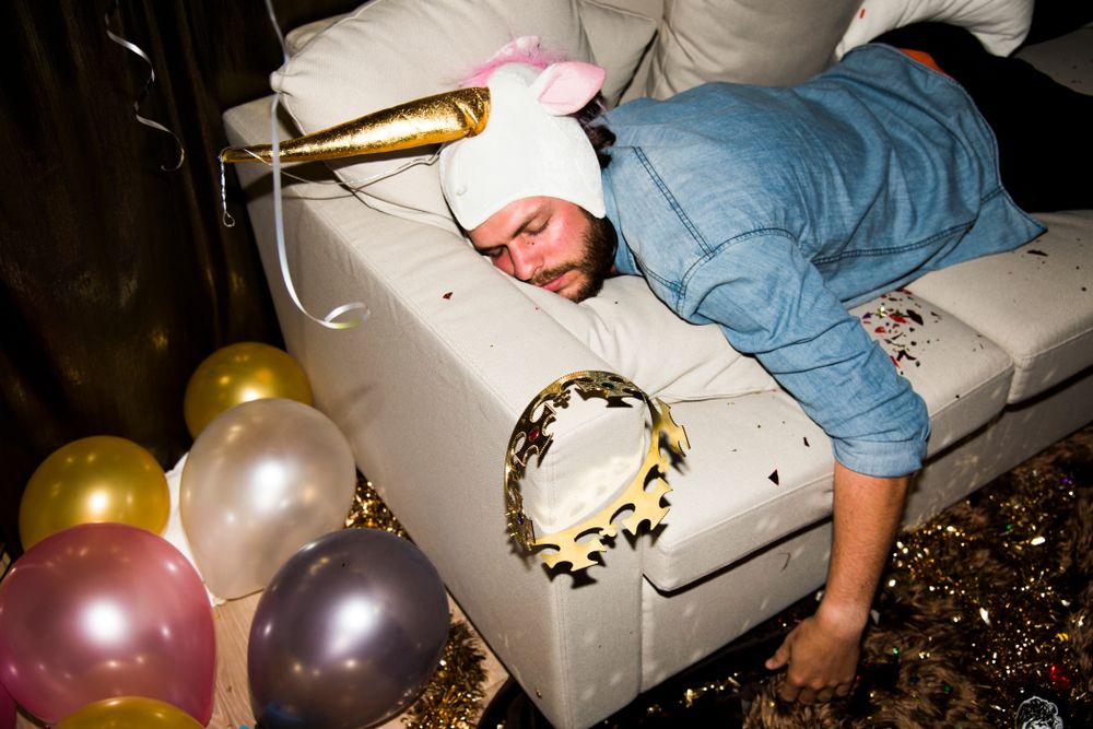 Man asleep on couch wearing unicorn hat, surrounded by balloons and party decorations.
