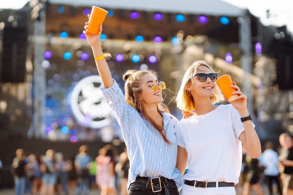Two young people at an outdoor music festival, holding orange cups, smiling in front of a stage with lights.