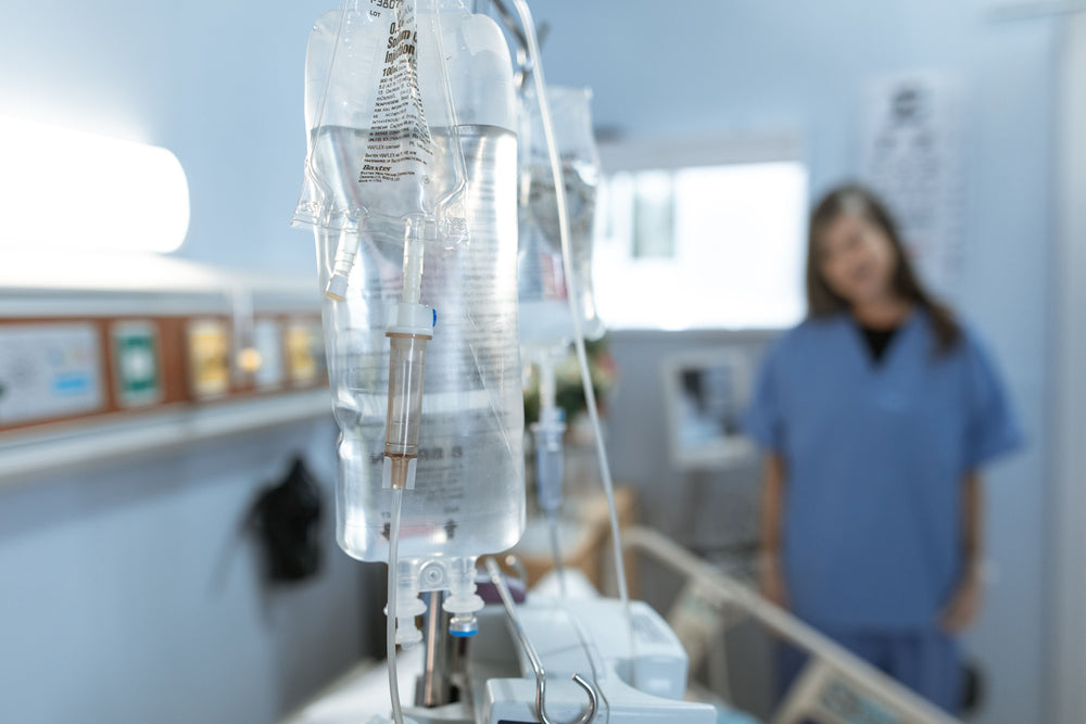 IV drip bag in hospital room; nurse in blue scrubs stands near bed.