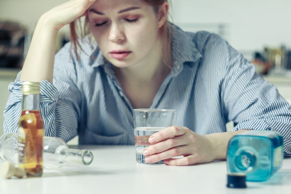 Woman with head in hand, holding water glass, bottles on table, appearing unwell.