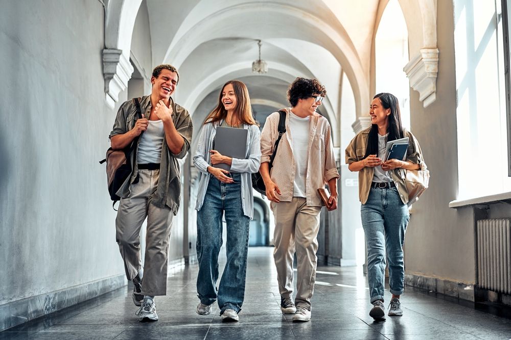 Four students walking and talking in a light-filled arched hallway; one carries a backpack, another a tablet.