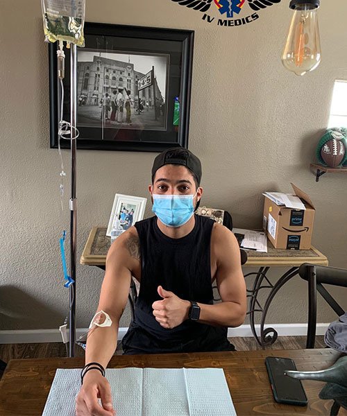 Man receiving IV fluids at home, wearing a mask and giving a thumbs-up.