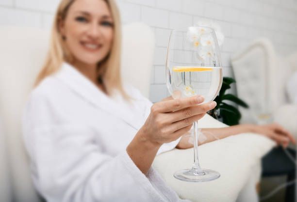 Woman in white robe smiling, holding a glass of water with lemon slice.