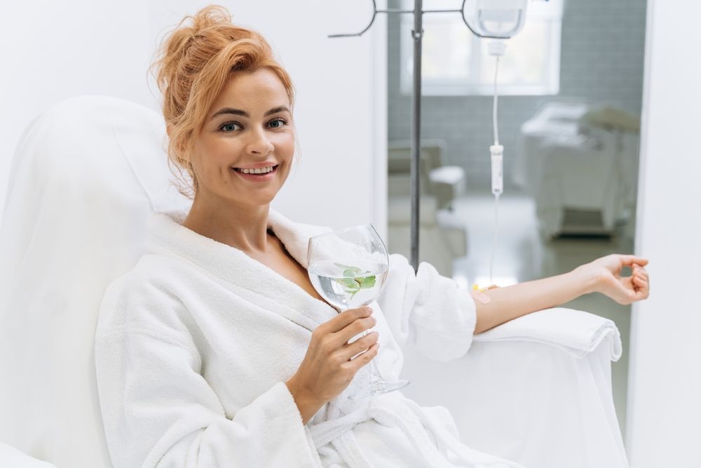 Woman in white robe, smiling, holding a drink, receiving IV drip in a bright room.