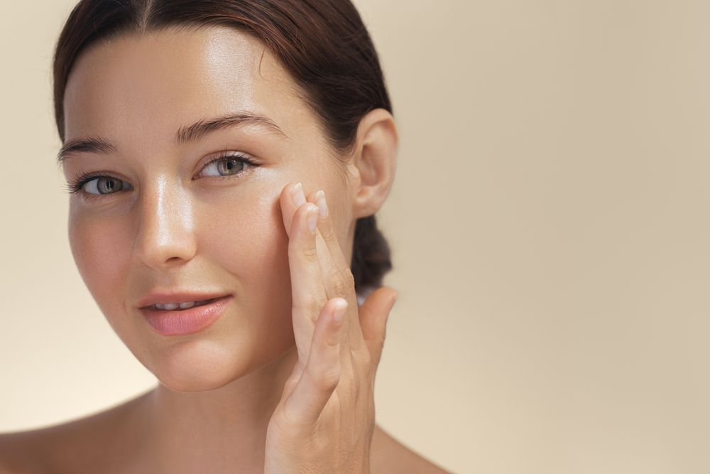 Woman with fair skin touches her face, looking toward the viewer against a beige backdrop.