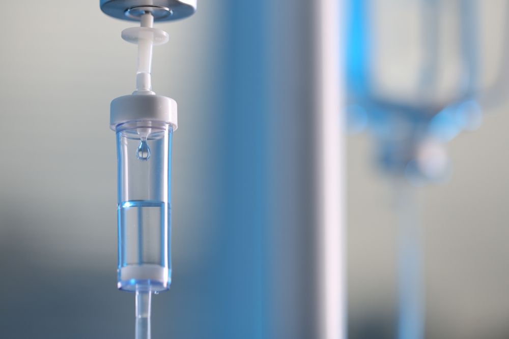 IV drip bag, hanging from a pole. Clear fluid dripping into a tube. Lit with blue light.