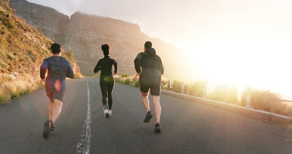 Three people running on a road with a mountain backdrop, sunlit sky.