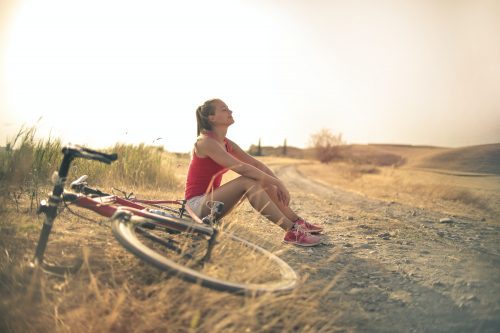 Woman in red tank and white shorts rests next to a bicycle on a dirt road, looking up at the sun.