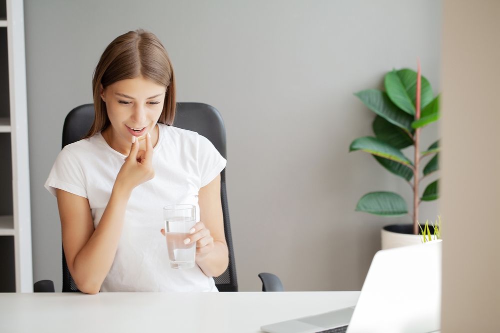 Woman taking a pill with a glass of water, sitting at a desk with a laptop and plant in the background.