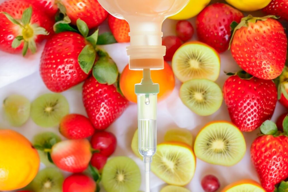 IV drip with liquid over assorted fruit: strawberries, kiwi, oranges, and grapes.