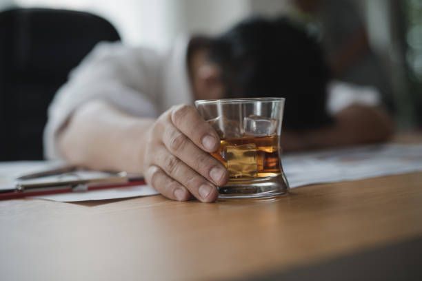 Person slumped over desk, hand outstretched to a glass of amber liquid.