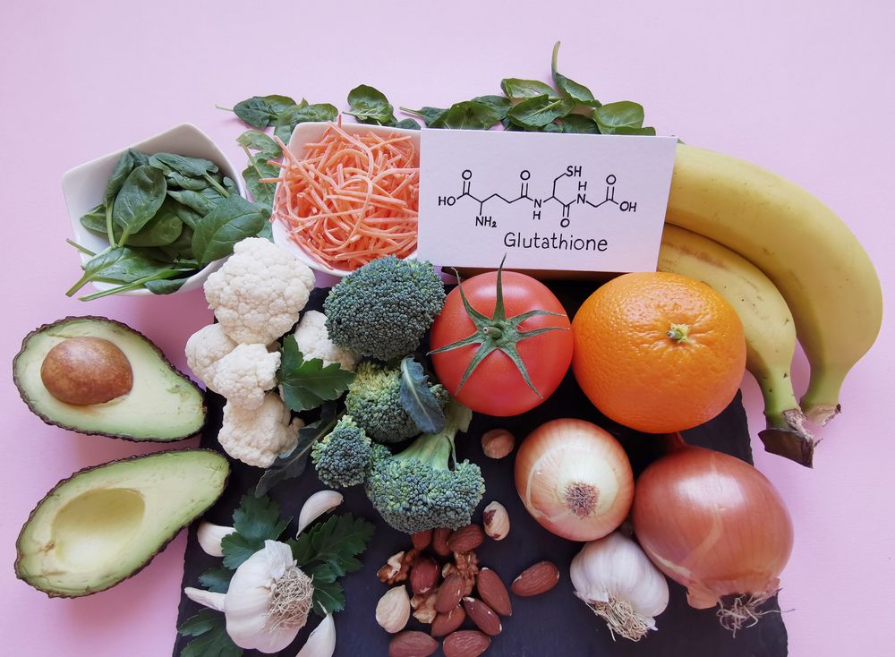 Assorted fruits and vegetables, including avocado, broccoli, and orange, surrounding a card with glutathione structure.