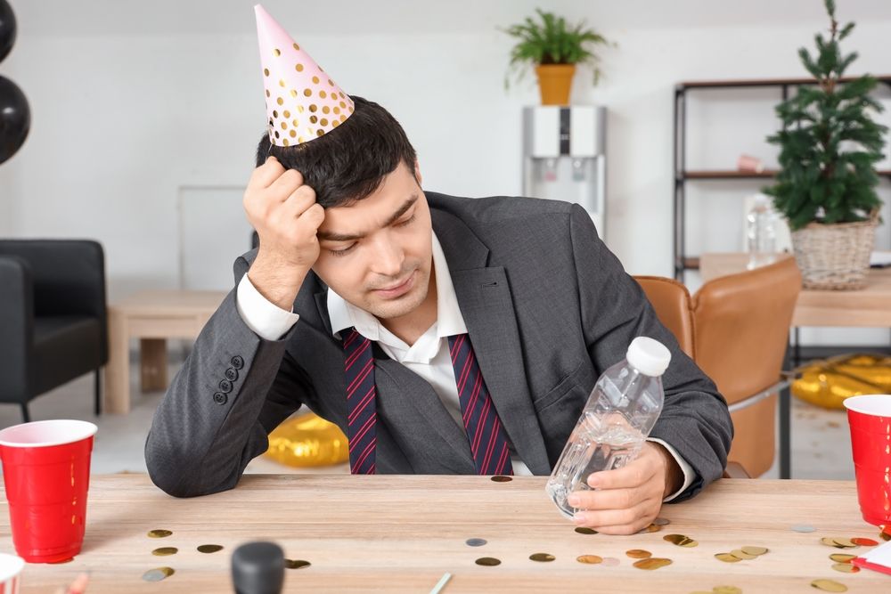 Man wearing party hat, slumped at desk, holding water bottle; office, scattered confetti.