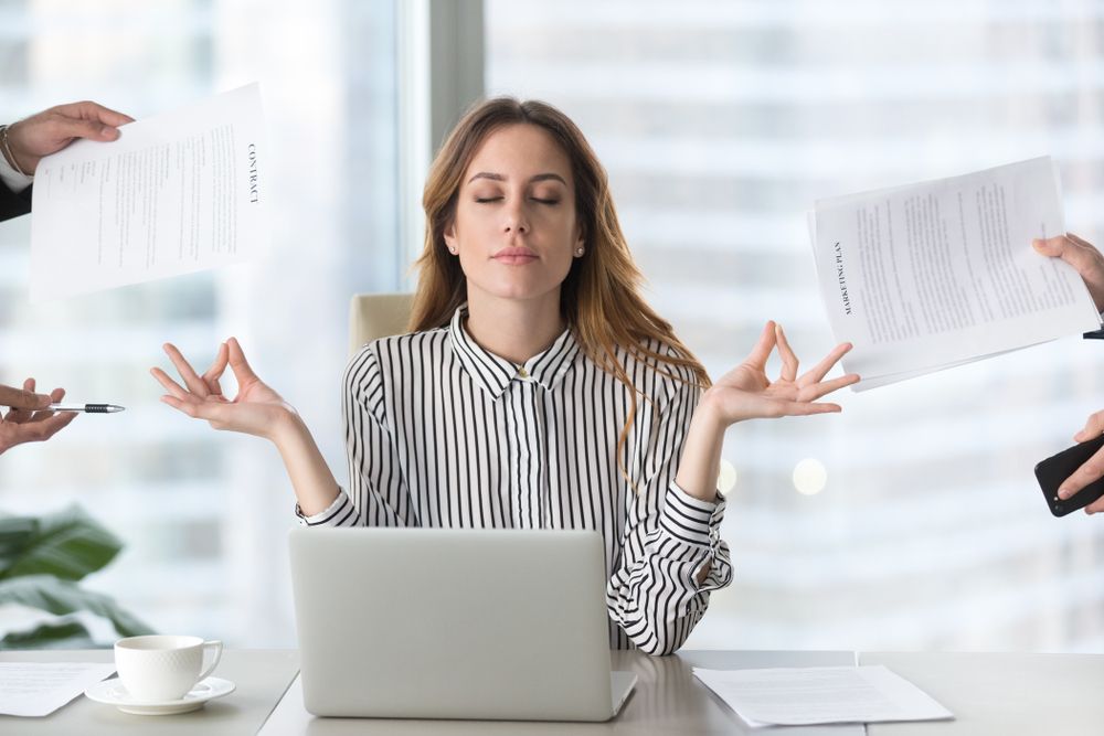 Woman meditating at desk with laptop and papers; arms outstretched, eyes closed.