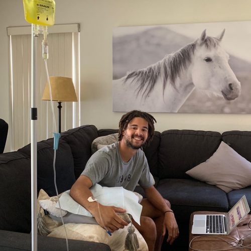 Man seated on a couch receiving an IV drip, smiling, with a laptop on a table. White horse artwork in the background.