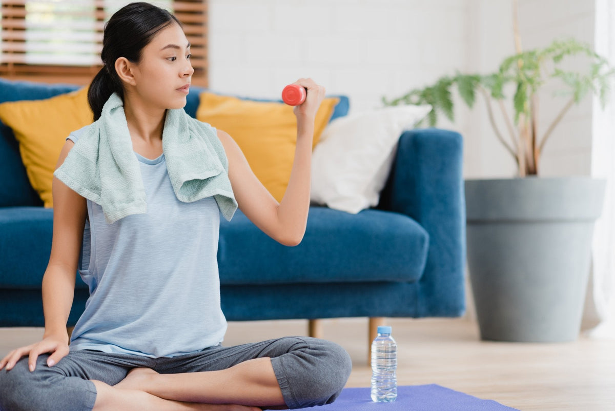 Woman exercising at home, lifting a dumbbell, seated on a yoga mat. Towel around neck, water bottle nearby.